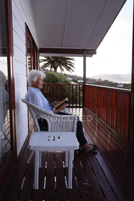 Вид сбоку активной пожилой женщины, читающей книгу, сидя дома на кресле на балконе — стоковое фото