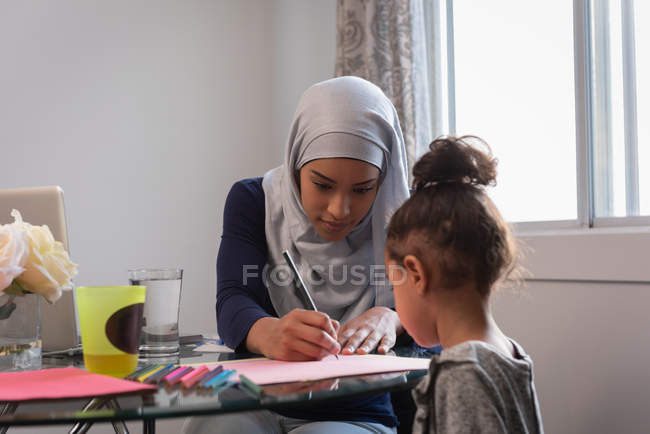 Visão frontal da mãe de raça mista vestindo hijab ensinando sua filha em casa em torno de uma mesa — Fotografia de Stock