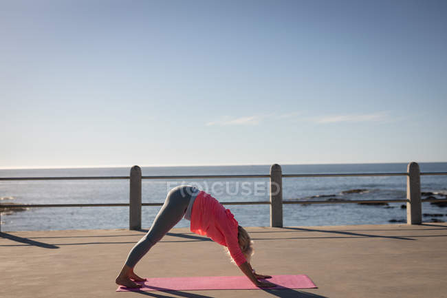 Rear view of an active senior woman practicing fitness exercises on a fitness mat on a promenade in the sunshine in front of the beach — Stock Photo