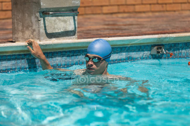 Vista frontal do jovem nadador caucasiano do sexo masculino agarrado à borda da piscina exterior enquanto usa touca de natação e óculos no dia ensolarado — Fotografia de Stock