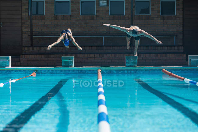 Вид спереди на мужчин и женщин, прыгающих в воду в одно и то же время в бассейне на солнце — стоковое фото