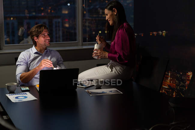 Вид спереду щасливих молодих багатоетнічних керівників офісів розмовляють один з одним, маючи їжу за столом в сучасному офісі — стокове фото