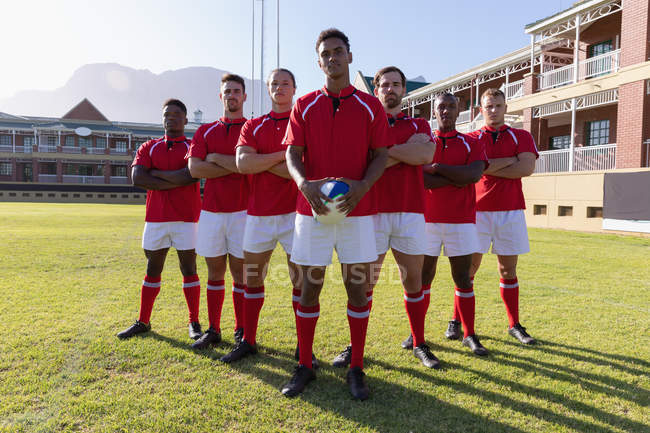 Vue de face d'une équipe de joueurs de rugby multiethniques masculins debout avec une balle de rugby sur le terrain de rugby par une journée ensoleillée — Photo de stock