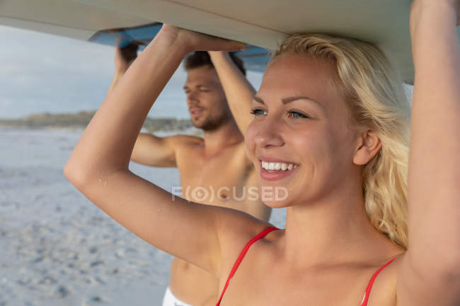 Vista lateral do casal carregando prancha de surf na praia no dia ensolarado. Estão a ver as ondas. — Fotografia de Stock