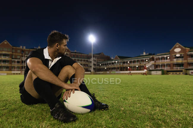 Vista frontal de um jogador de rugby masculino chateado sentado com uma bola de rugby no estádio à noite — Fotografia de Stock