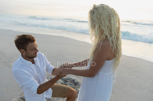 Vista lateral del hombre guapo cogido de la mano de la mujer rubia en la playa. Él le pregunta en el compromiso - foto de stock