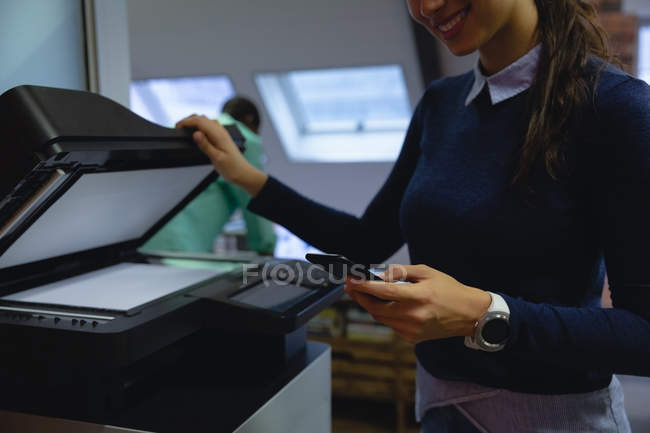 Sección media de la mujer de negocios sonriente usando el teléfono móvil mientras sostiene la fotocopiadora xerox en la mano en la oficina - foto de stock