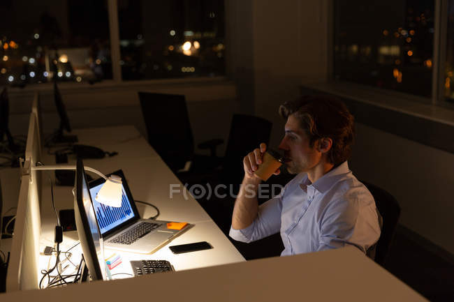 Вид збоку красивого молодого чоловіка-виконавця п'є каву, працюючи за столом в сучасному офісі. Він працює пізно — стокове фото