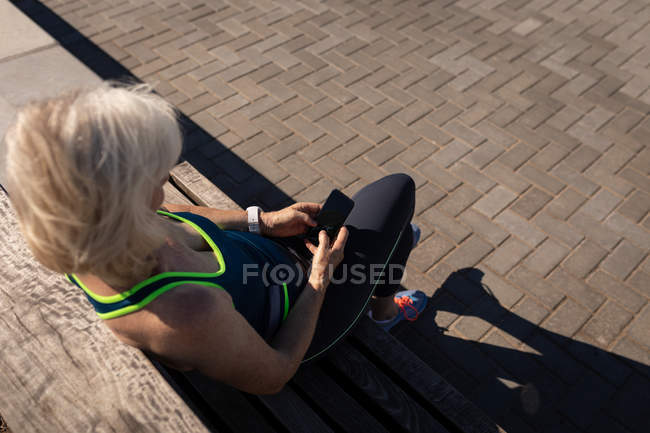Visão de alto ângulo de uma mulher idosa ativa usando seu telefone celular e sentada em um banco em um passeio sob a luz do sol — Fotografia de Stock