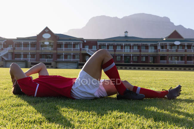 Vista lateral de um jogador de rugby caucasiano machucado deitado no chão ao sol — Fotografia de Stock