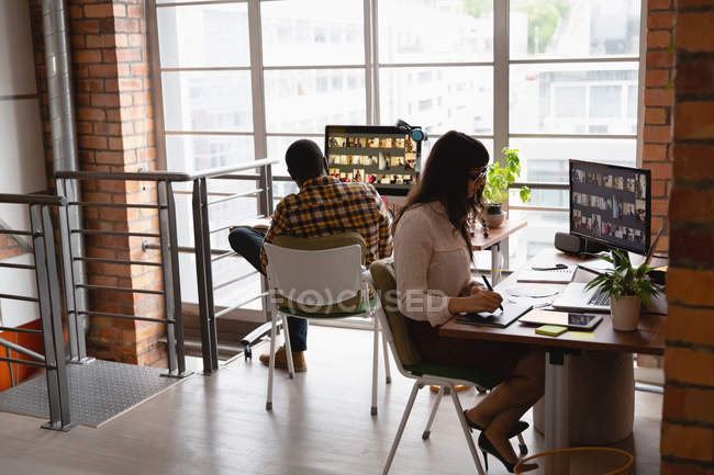 Вид сбоку на кавказскую деловую женщину, работающую за графическим планшетом, в то время как афроамериканец, работающий за ней в офисе — стоковое фото