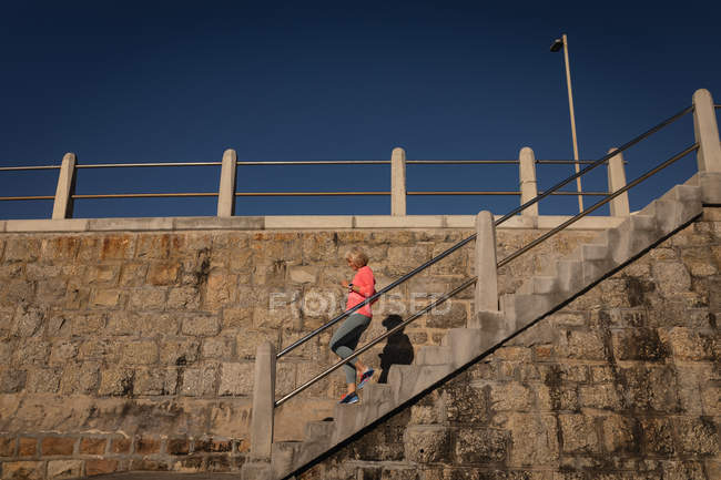 Низький кут зору активної старшої жінки, що йде по сходах на набережній поруч з пляжем під сонцем — стокове фото