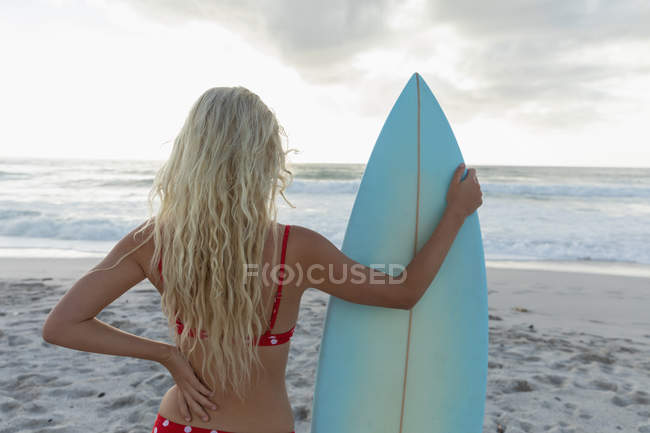 Vista traseira do surfista feminino com uma prancha de surf em pé em uma praia em um dia ensolarado. Ela está a ver as ondas. — Fotografia de Stock