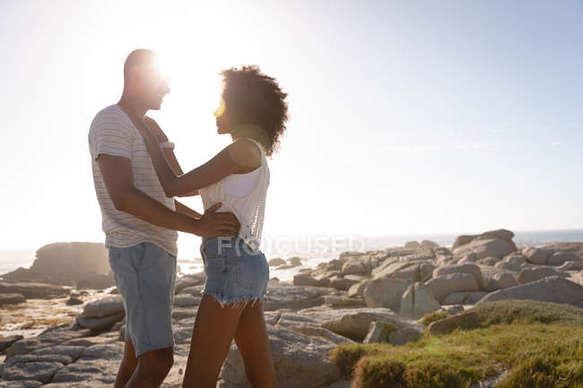 Vista laterale della coppia afro-americana che si abbraccia in piedi vicino al mare sulla roccia — Foto stock