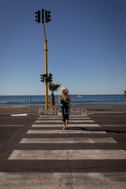 Vue de face d'une femme âgée active faisant du jogging au croisement piétonnier d'une route au bord de la mer sous le soleil — Photo de stock