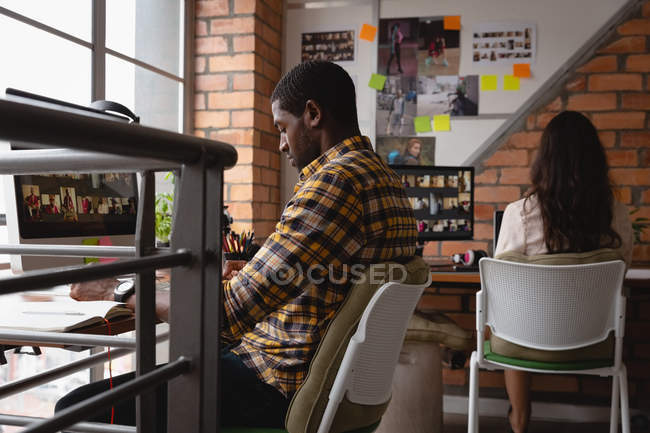 Vue latérale de l'homme d'affaires afro-américain travaillant sur ordinateur portable tandis que la femme caucasienne travaillant derrière lui au bureau — Photo de stock