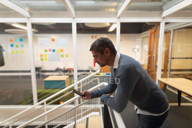 Side view of a Caucasian businessman using his mobile phone in office while leaning on the security barriers — Stock Photo