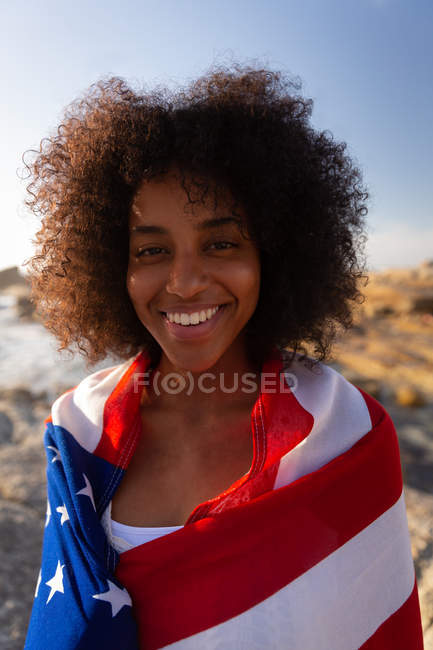 Vue de face de la femme afro-américaine debout et enveloppé drapeau américain près du bord de la mer tout en regardant la caméra — Photo de stock
