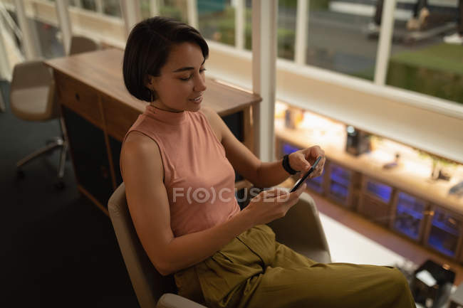 Visão de alto ângulo de uma empresária asiática usando seu telefone celular enquanto sentada no escritório — Fotografia de Stock