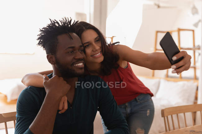 Vista frontal do casal multi étnico sorrindo e tirando selfie em casa — Fotografia de Stock