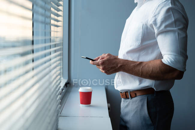 Средняя секция молодого мужчины, использующего мобильный телефон, глядя в окно современного офиса — стоковое фото