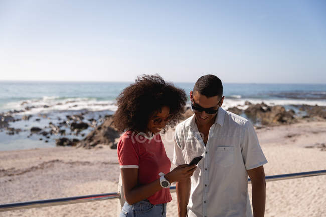Frontansicht eines afrikanisch-amerikanischen Paares, das sein Handy benutzt und an einem sonnigen Tag am Strand lächelt — Stockfoto
