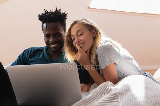 Vista frontal do casal multi étnico sorrindo, sentado e usando laptop em casa no sofá — Fotografia de Stock