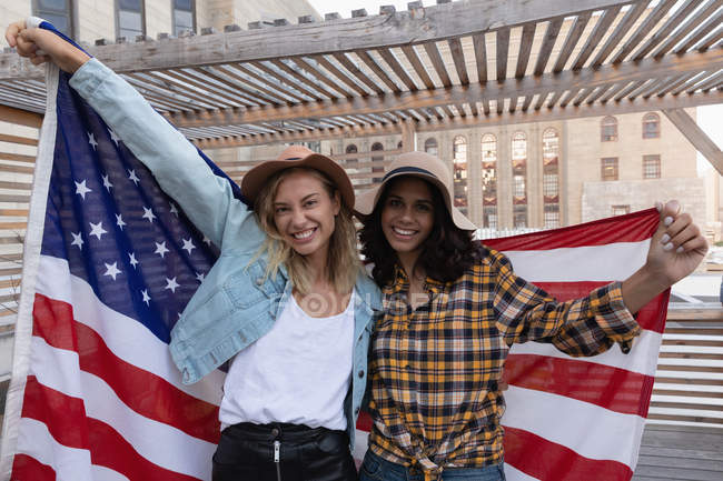 Vista frontal de múltiples amigas étnicas con bandera americana en el balcón en casa - foto de stock
