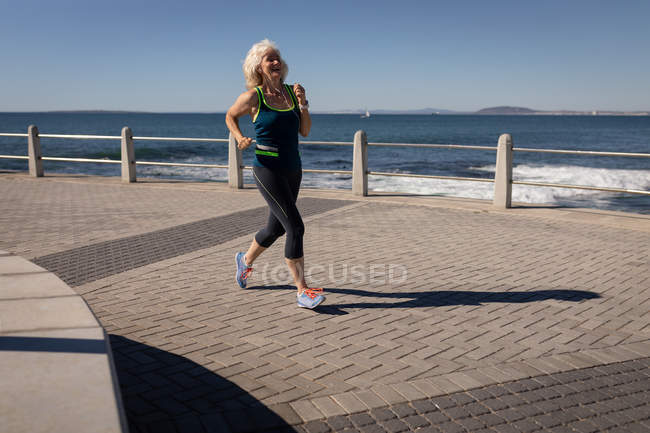 Vista frontal de uma mulher idosa ativa fazendo jogging em um passeio ao longo do mar sob o sol — Fotografia de Stock