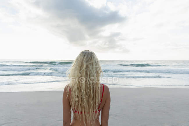 Вид сзади на красивую женщину, стоящую на пляже в солнечный день. Она смотрит на океан. — стоковое фото