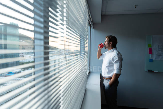 Side view of young male executive with hand in pocket drinking coffee while looking outside in a modern office — Stock Photo