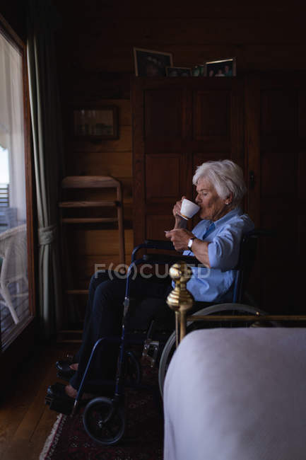 Side view of a disabled active senior woman drinking a coffee and sitting on a wheelchair in bedroom at home — Stock Photo