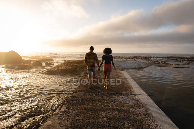 Vista trasera de la pareja afroamericana caminando y relajándose en el lado del mar. Están tomados de la mano. - foto de stock