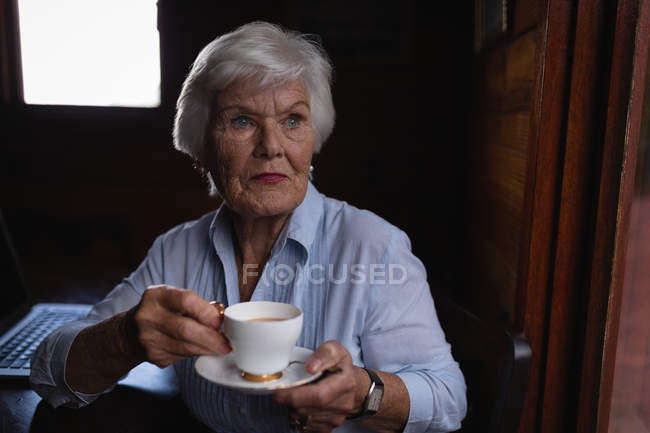 Vue de face d'une femme âgée active réfléchie regardant ailleurs tout en prenant un café à la maison — Photo de stock