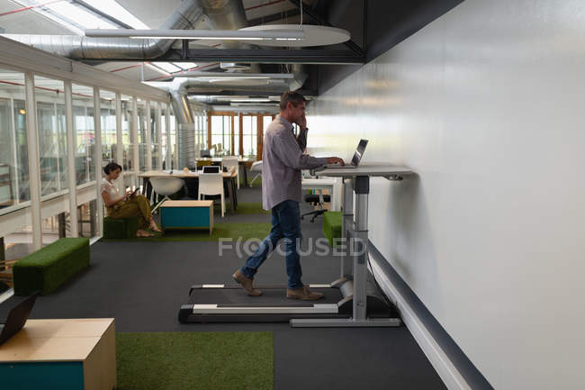 Vista lateral de un hombre de negocios caucásico que trabaja en un ordenador portátil y habla en su teléfono móvil mientras hace ejercicio en una cinta de correr en la oficina - foto de stock