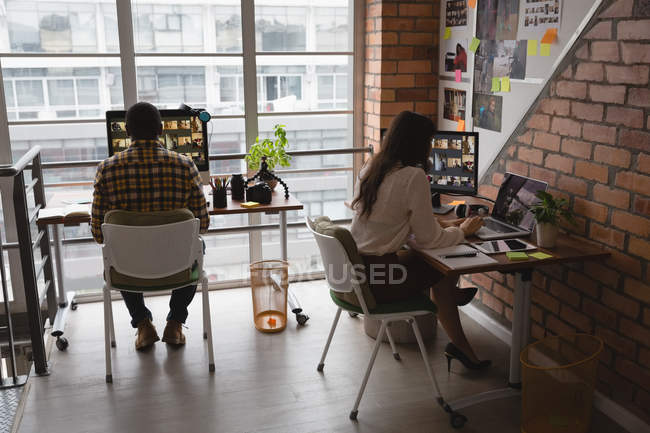 Задний вид разнообразных деловых коллег, просматривающих фотографии на ноутбуках и настольных компьютерах во время работы в современном офисе — стоковое фото