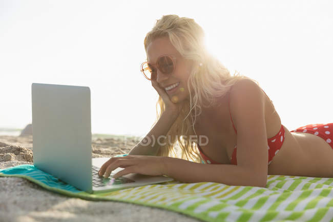 Vista lateral de la mujer usando el ordenador portátil mientras está acostado en la playa en un día soleado. Ella es feliz. - foto de stock