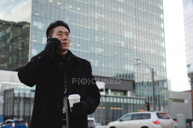 Seitenansicht eines jungen asiatischen männlichen Geschäftsmannes, der auf dem Handy spricht, während er einen Kaffee in der Hand hält und hinter sich ein Gebäude in der Stadt baut — Stockfoto