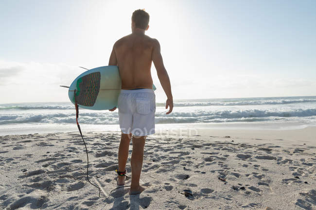 Jeune surfeur masculin avec une planche de surf debout sur une plage par une journée ensoleillée. Il regarde les vagues — Photo de stock