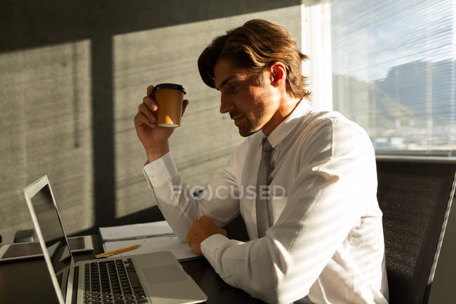 Side view of handsome young male executive having coffee while working on laptop at table in a modern office — Stock Photo