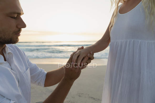 Вид сбоку на красивого мужчину, надевающего кольцо женщине в палец на пляже. Он просит её о помолвке. — стоковое фото