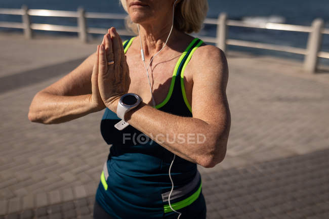 Sección media de una mujer mayor activa realizando yoga en un paseo marítimo bajo el sol - foto de stock