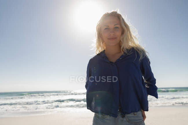 Retrato de una hermosa mujer rubia de pie en la playa en un día soleado. Ella está mirando y sonriendo a la cámara - foto de stock