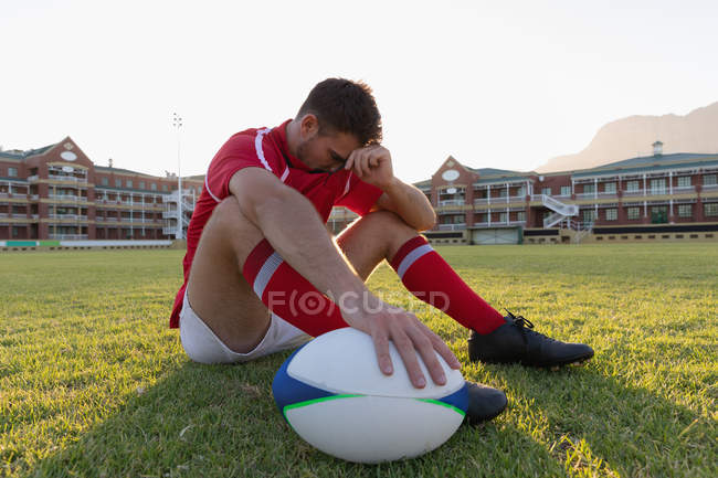 Seitenansicht eines aufgebrachten kaukasischen Rugbyspielers, der mit Rugbyball im Stadion sitzt und seinen Kopf hält — Stockfoto