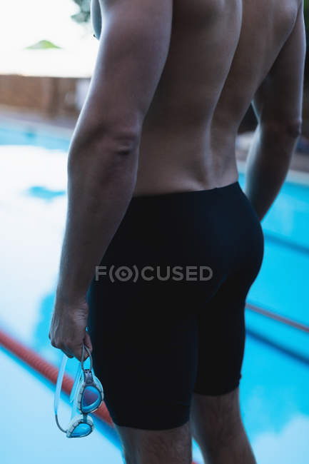 Seção intermediária de um nadador masculino em pé em frente à piscina com seu óculos de natação na mão — Fotografia de Stock