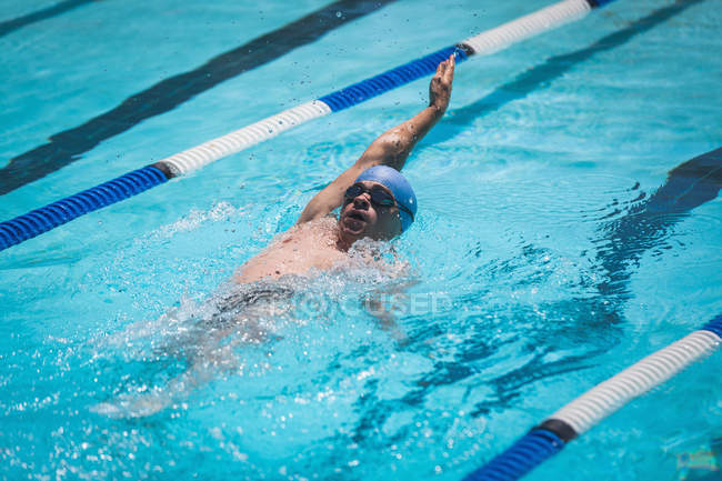 Vista frontale del giovane nuotatore maschio caucasico che nuota dorso nella piscina all'aperto sotto il sole — Foto stock