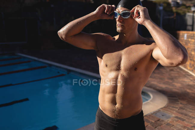Vista frontal de un nadador caucásico masculino con sus gafas de natación mientras está de pie cerca de la piscina en un día soleado - foto de stock