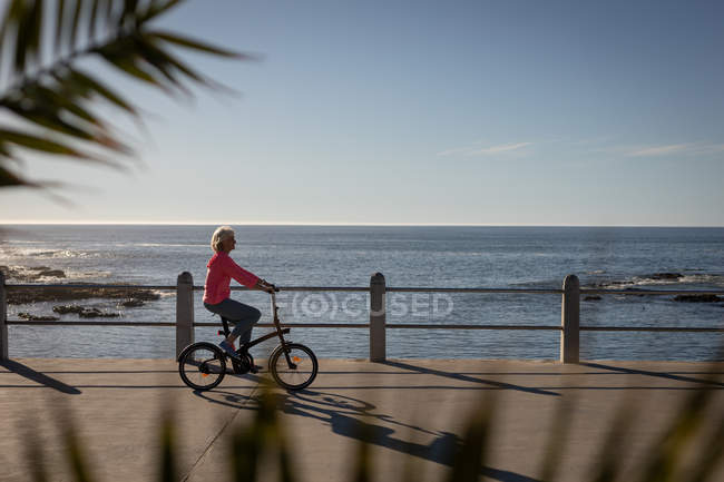 Side view of an active senior woman riding bicycle on a promenade along the beach on the evening — Stock Photo