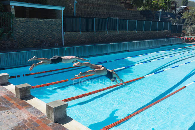 Vista de ángulo alto de nadadores caucásicos masculinos y femeninos saltando al agua al mismo tiempo en la piscina bajo el sol - foto de stock