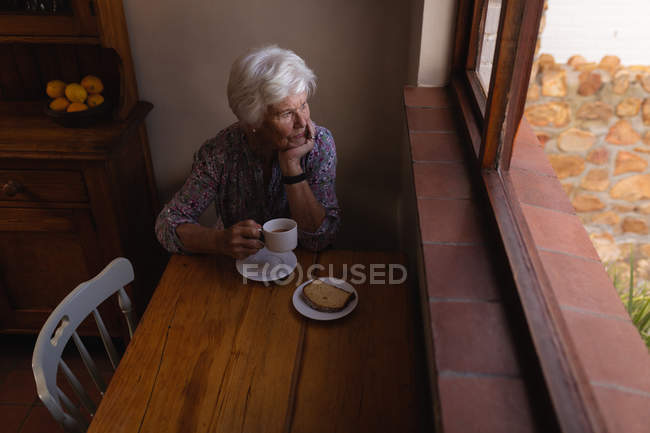 Vista de alto ângulo de uma mulher sênior ativa pensativa olhando pela janela enquanto toma café na cozinha em casa — Fotografia de Stock